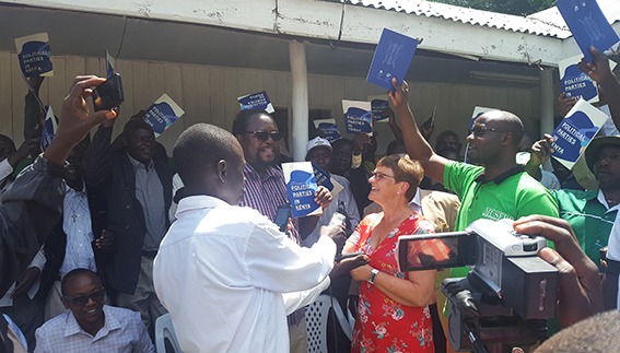 Trude Brosvik har mange kenyanske mennesker rundt seg. Noen vifter med en blå håndbok. En filmer.