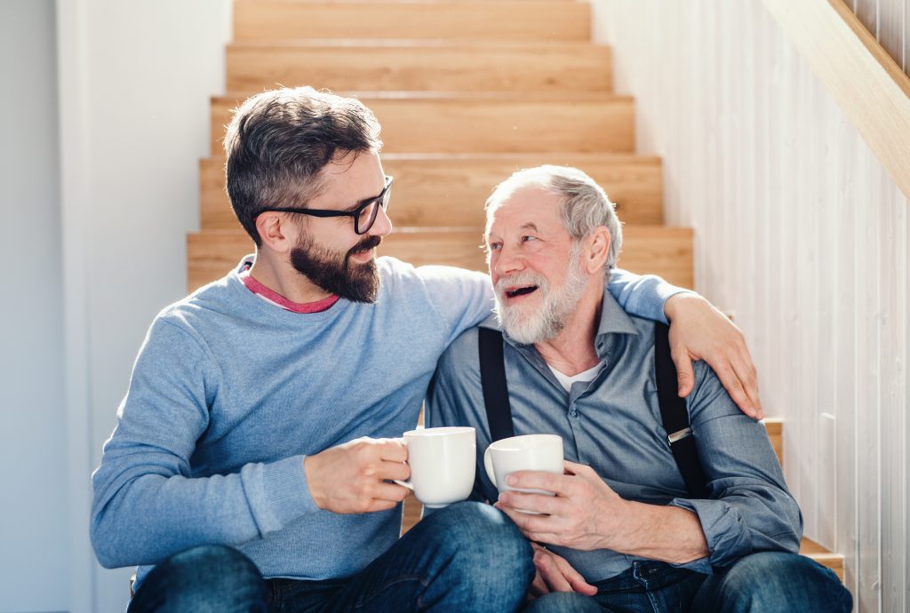 Eldre mann smiler og klemmer sin sønn. De to drikker en kopp kaffe, har på seg blå klær og sitter i en trapp.