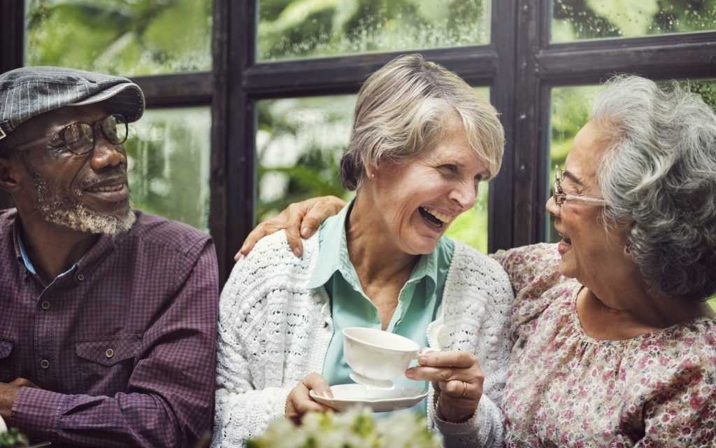 Tre eldre venner drikker kaffe og smiler til hverandre i et drivhus