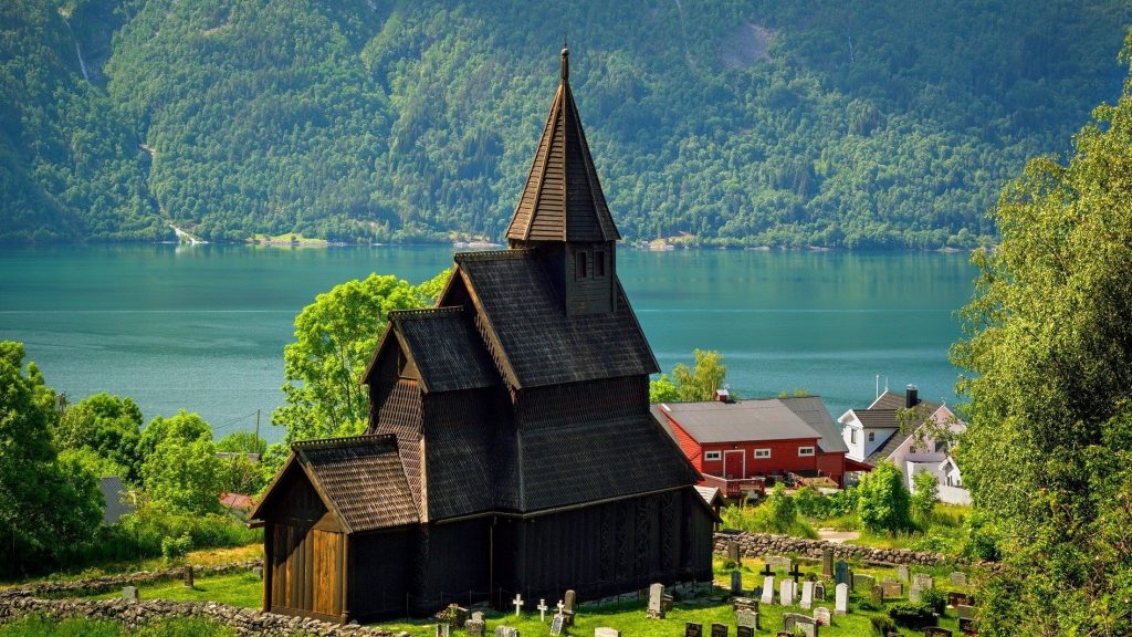 En norsk stavkirke står plassert høyt over en fjord. Blå vann, grønt gress og brun kirke