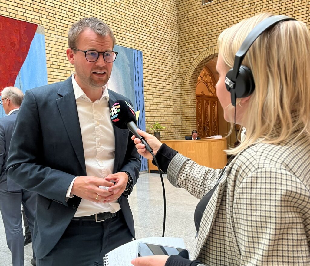 Kjell Ingolf står i vandrehallen og blir intervjuet av en journalist. Han snakker inn i en mikrofon